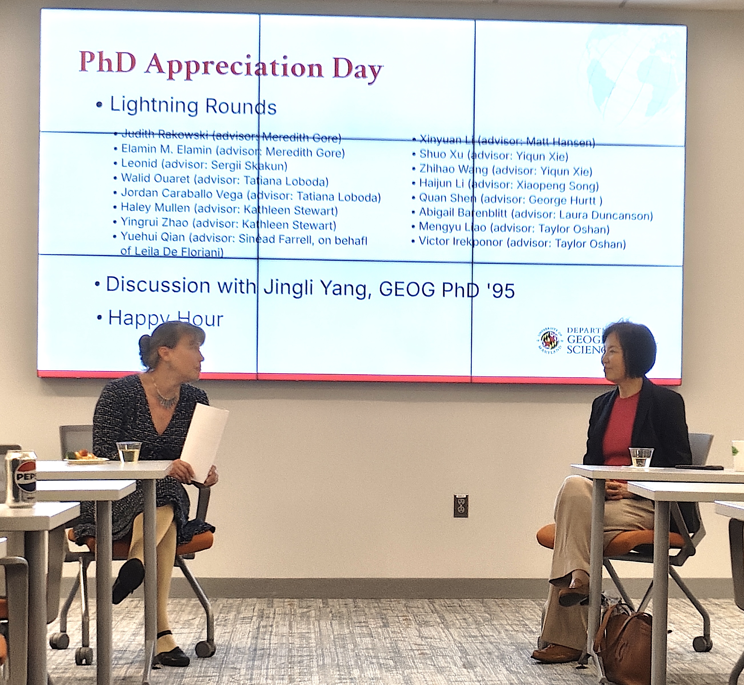 A professor in a dark patterned dress and a woman in a black blazer and pink top sit in chairs facing each other, engaged in a discussion. Behind them, a large digital screen displays a 