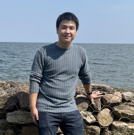 A young East Asian man smiling at the camera outdoors, leaning his hand against a wire-mesh rock wall along a body of water. He is wearing a long-sleeved ribbed grey sweater with pushed-up sleeves and dark blue jeans under a clear blue sky.