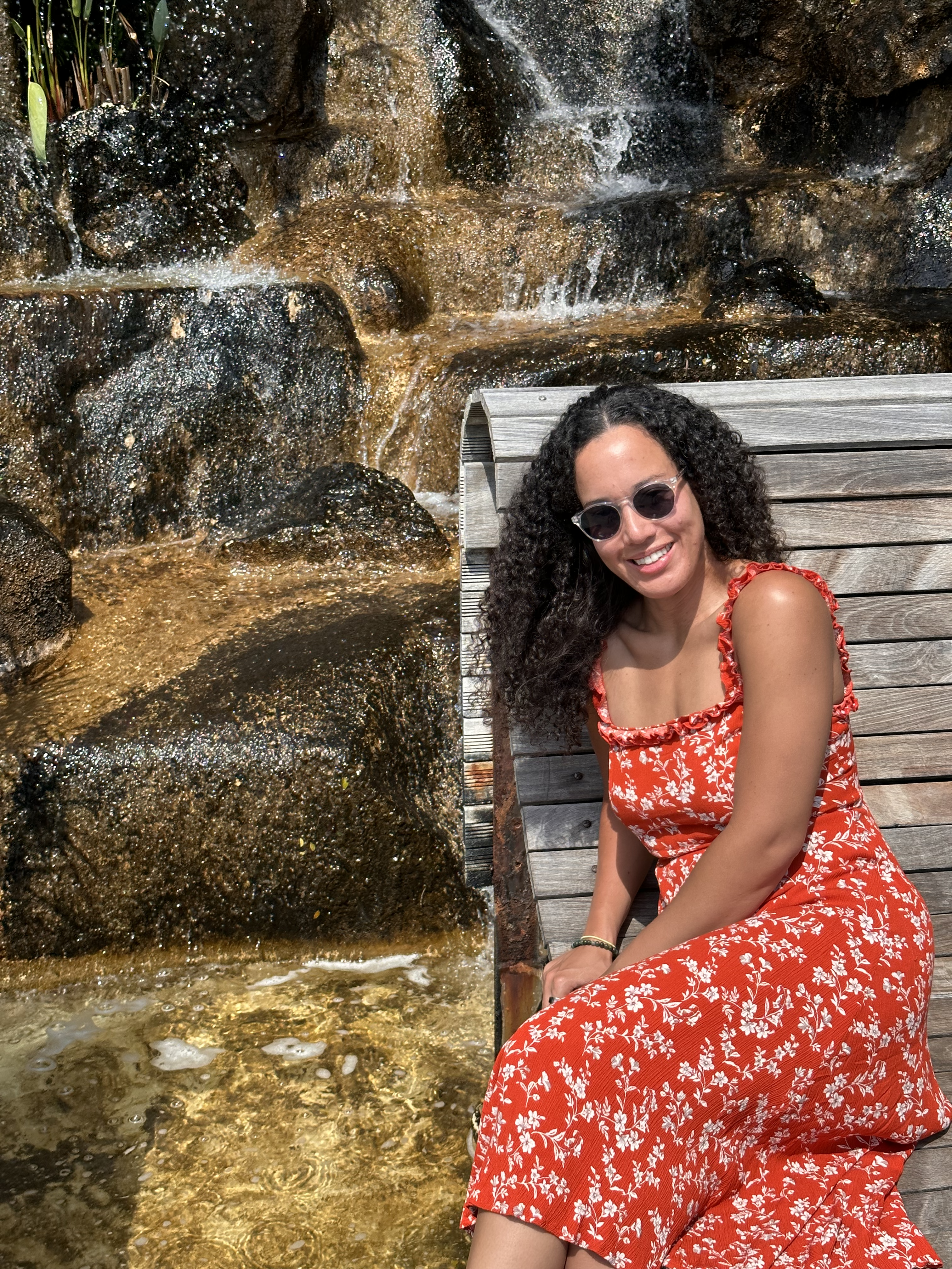 Kristen Halliday, wearing darc sits on a bench in front of waterfall. 