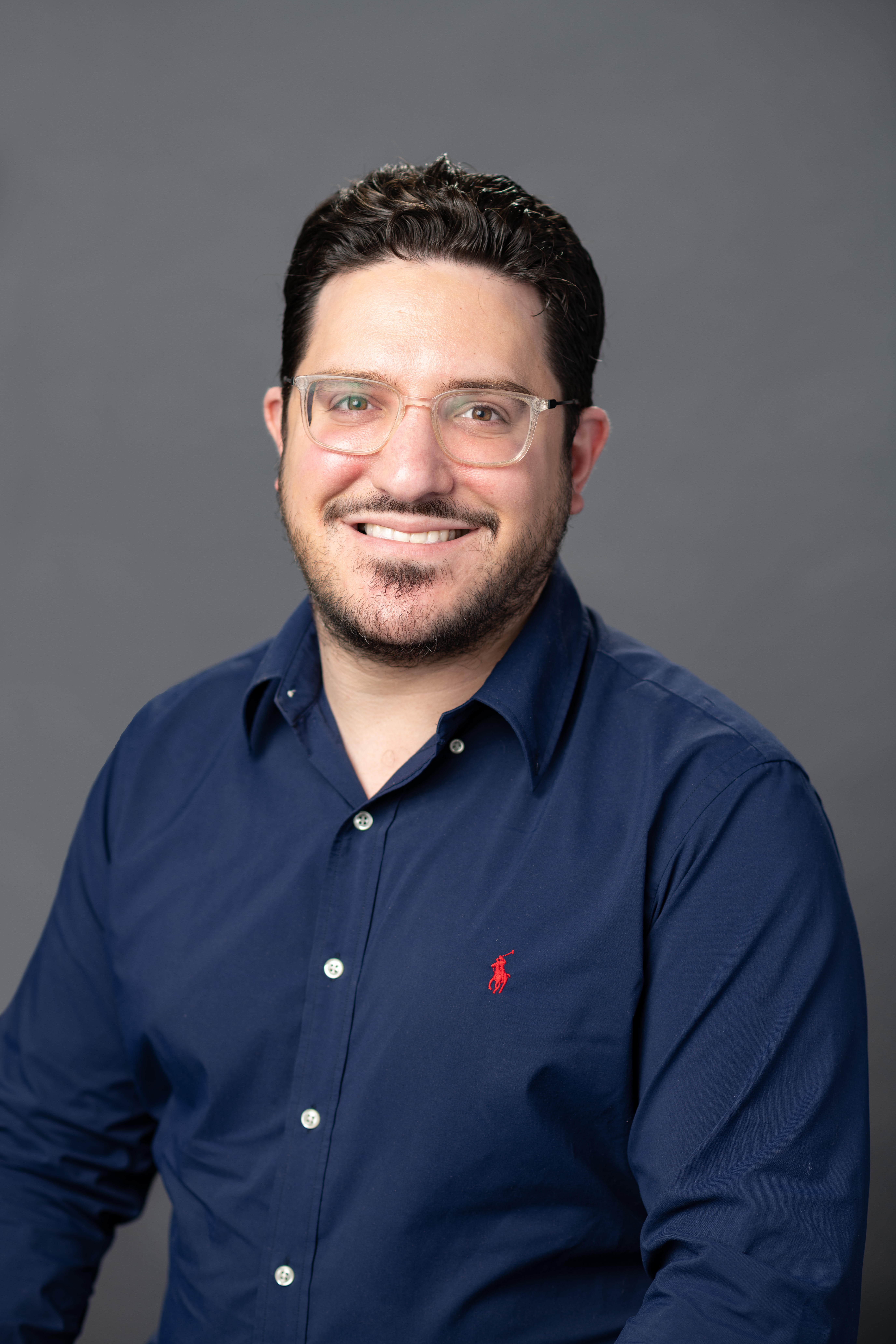 Aaron Goldman, wearing a blue shirt and glasses, smiles warmly at the camera.
