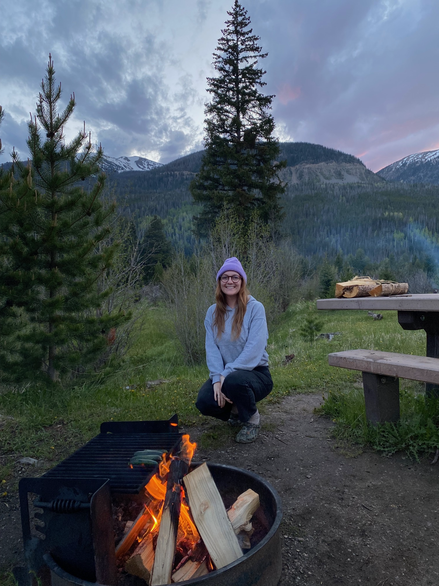 A woman enjoying a warm campfire, seated comfortably on the ground, with trees in the background.