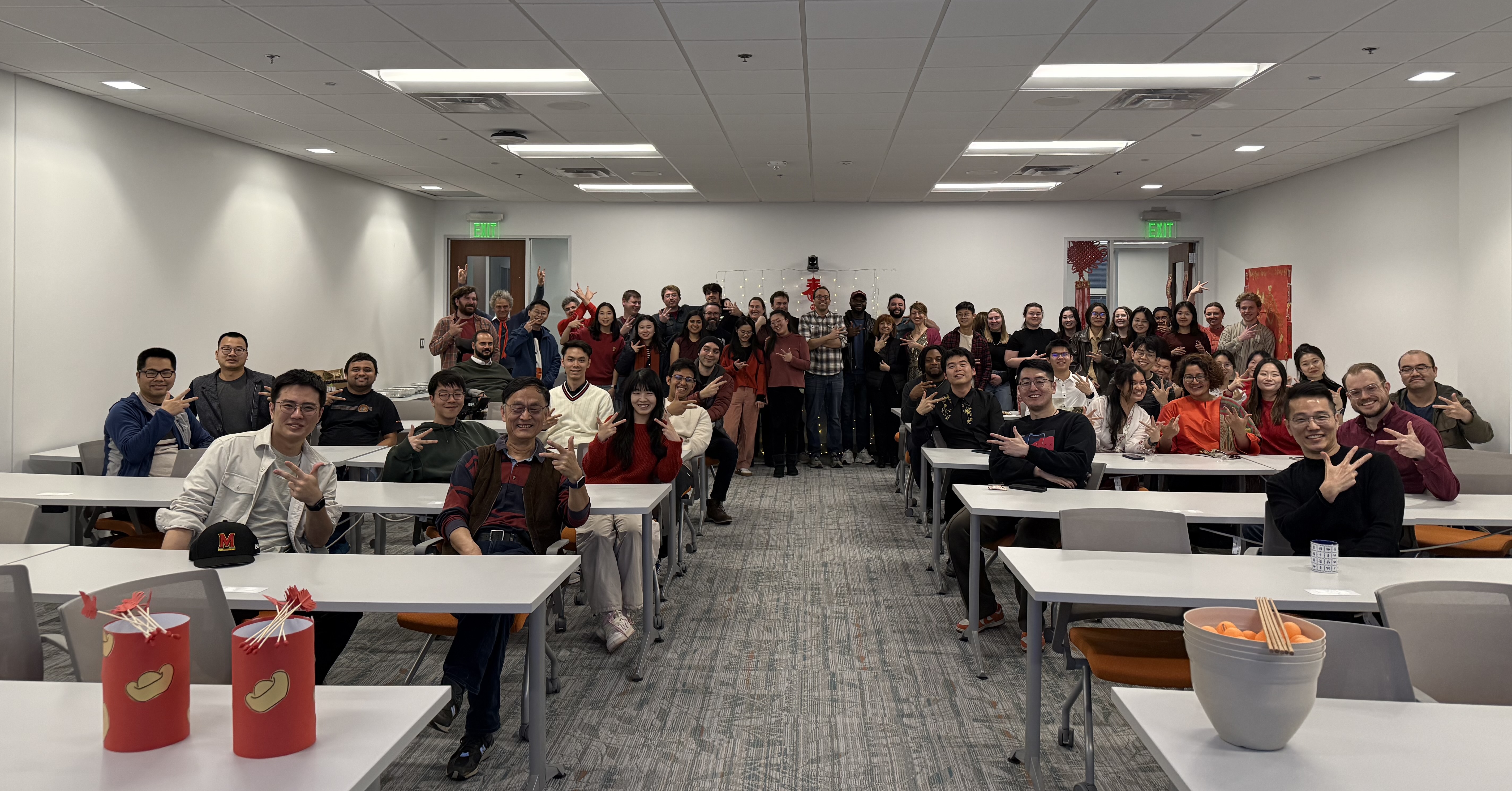 A large group of approximately 50 people of diverse backgrounds poses for a photo in a brightly lit modern office or classroom setting, many making a three-fingered hand sign representing innovation. The room is decorated for Lunar New Year with red banners, a