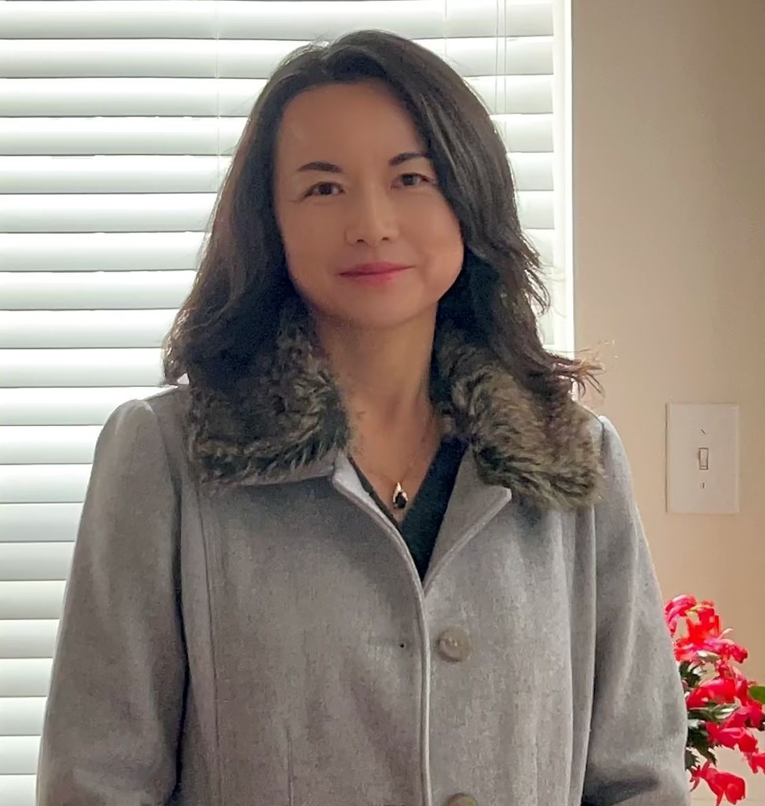 A woman with shoulder-length wavy dark hair smiling warmly at the camera. She is wearing a grey wool coat with a faux-fur collar and a small pendant necklace. She is standing indoors in front of a window with closed white blinds; a red plant is partially visible in the background.