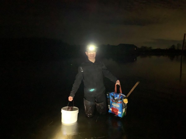 A man wearing a headlamp and chest waders stands knee-deep in a dark body of water at night. He's smiling and holding a white plastic bucket in one hand and a large, blue reusable shopping bag in the other. The headlamp illuminates the person and the immediate surface of the still water, while the background shows a dark shoreline under a dim, night sky.