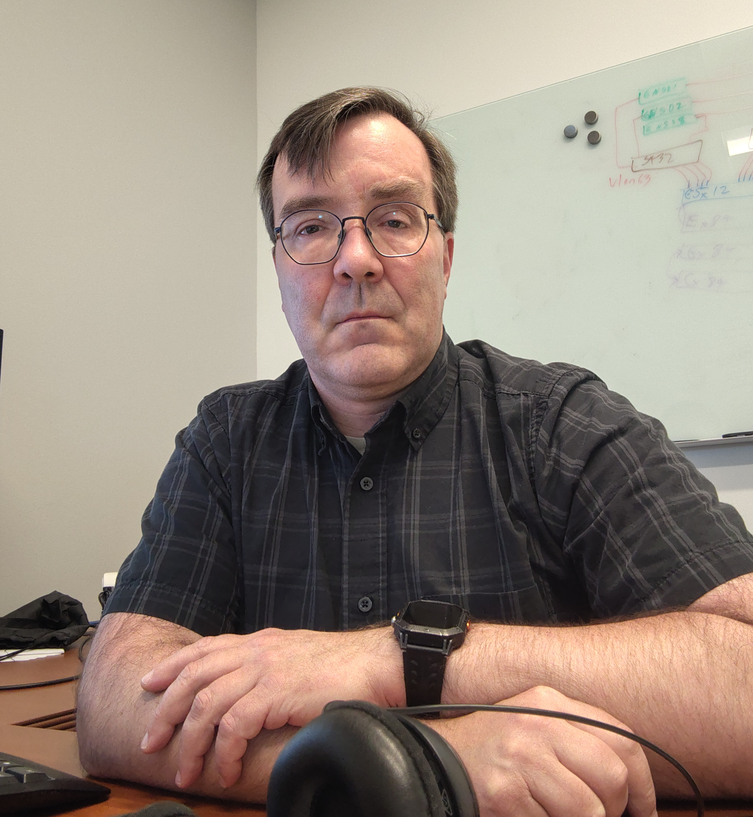Close-up portrait of a man with short brown hair and round glasses, wearing a dark plaid button-up shirt and a black smartwatch. He is sitting at a wooden desk with his arms crossed, looking forward with a neutral expression.