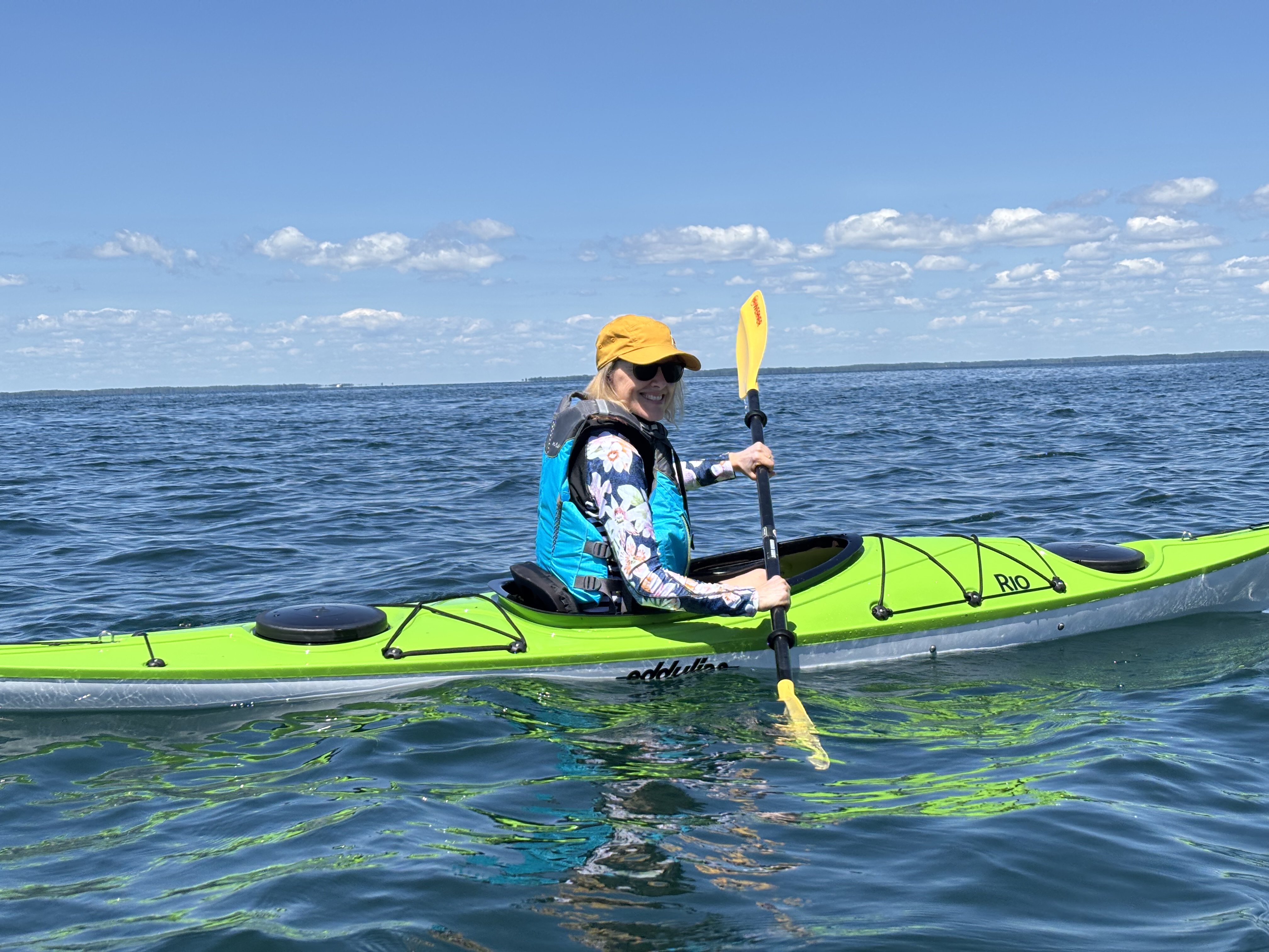 A person smiling while paddling a bright lime green kayak on calm, open blue water under a clear sunny sky.