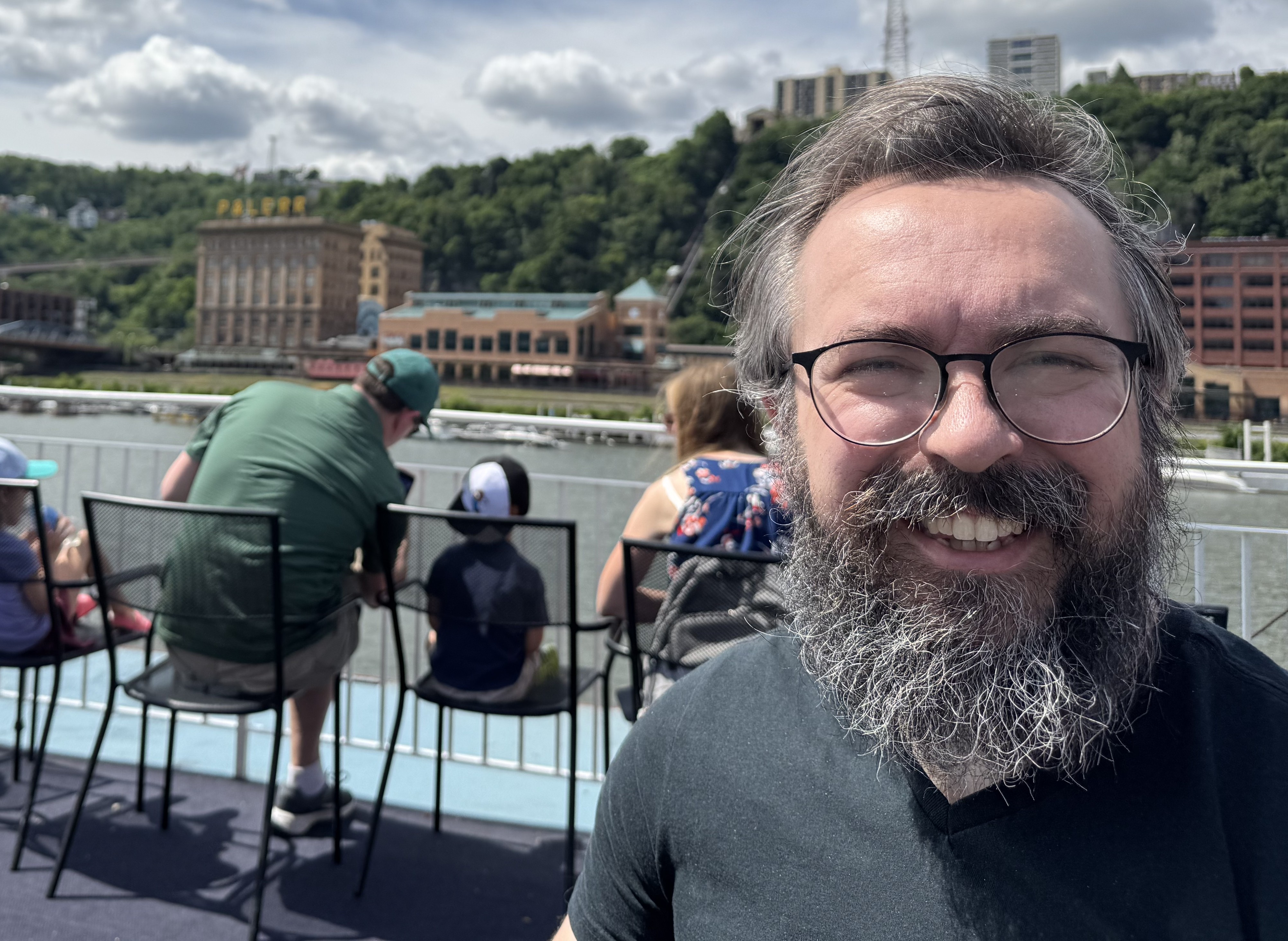 Close-up selfie of a smiling man with round glasses, a full graying beard, and a dark shirt on the outdoor deck of a boat. In the background, other passengers sit in chairs looking out toward a riverfront city skyline with buildings and a lush, tree-lined hill under a partly cloudy sky.