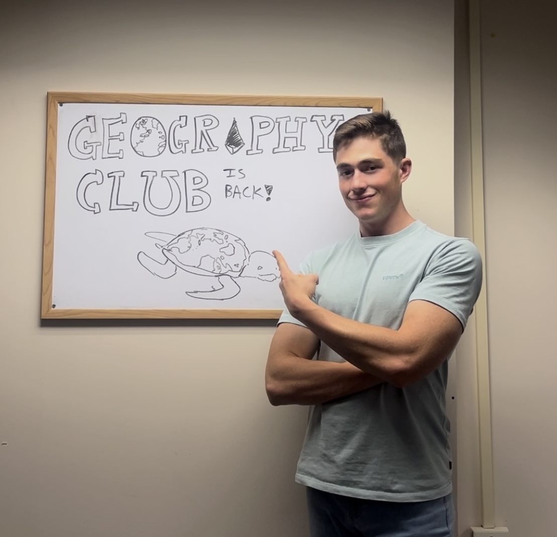 A young man in a light blue t-shirt stands next to a whiteboard framed on a plain wall. He has short brown hair, crossed arms, and points back over his shoulder toward the board with a slight smile. On the whiteboard, drawn in black marker, block letters spell out "GEOGRAPHY CLUB IS BACK!" with a globe integrated into the letter 'O' and an illustration of a sea turtle below the text whose shell resembles a world map.