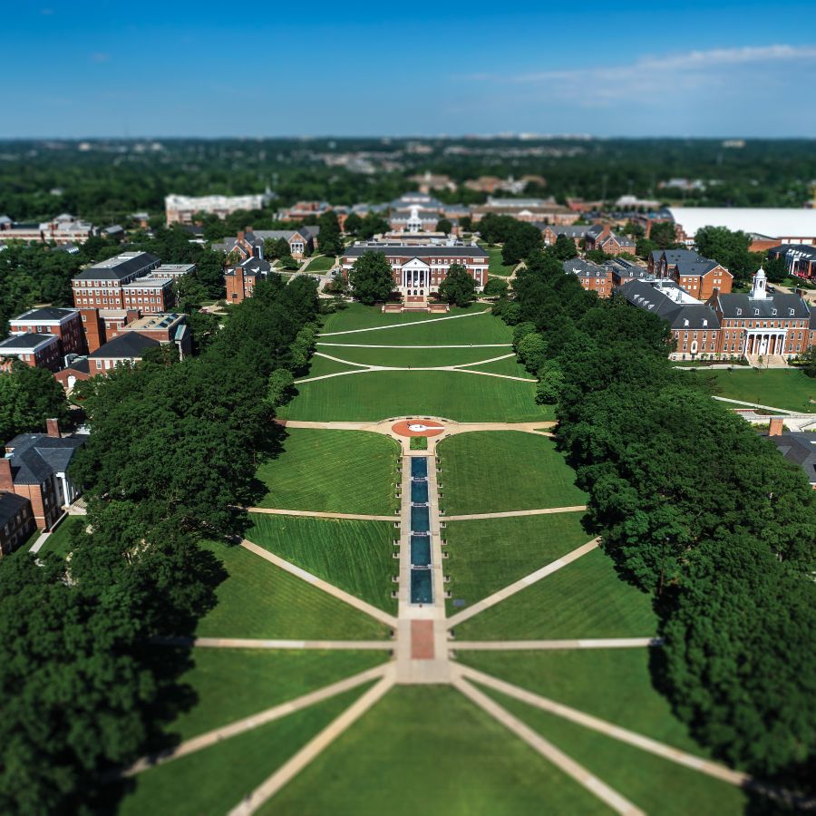 An aerial, tilt-shift photograph of McKeldin Mall at the University of Maryland, College Park. The shot looks down the long, rectangular green space toward McKeldin Library in the distance. Symmetrical paved pathways crisscross the vibrant green lawn, leading to the Omicron Delta Kappa (ODK) Fountain and a series of rectangular reflecting pools in the center. Rows of lush, mature trees line both sides of the mall, partially obscuring the red-brick academic buildings. The tilt-shift effect blurs the top and bottom of the frame, giving the campus a miniature, model-like appearance under a clear blue sky.