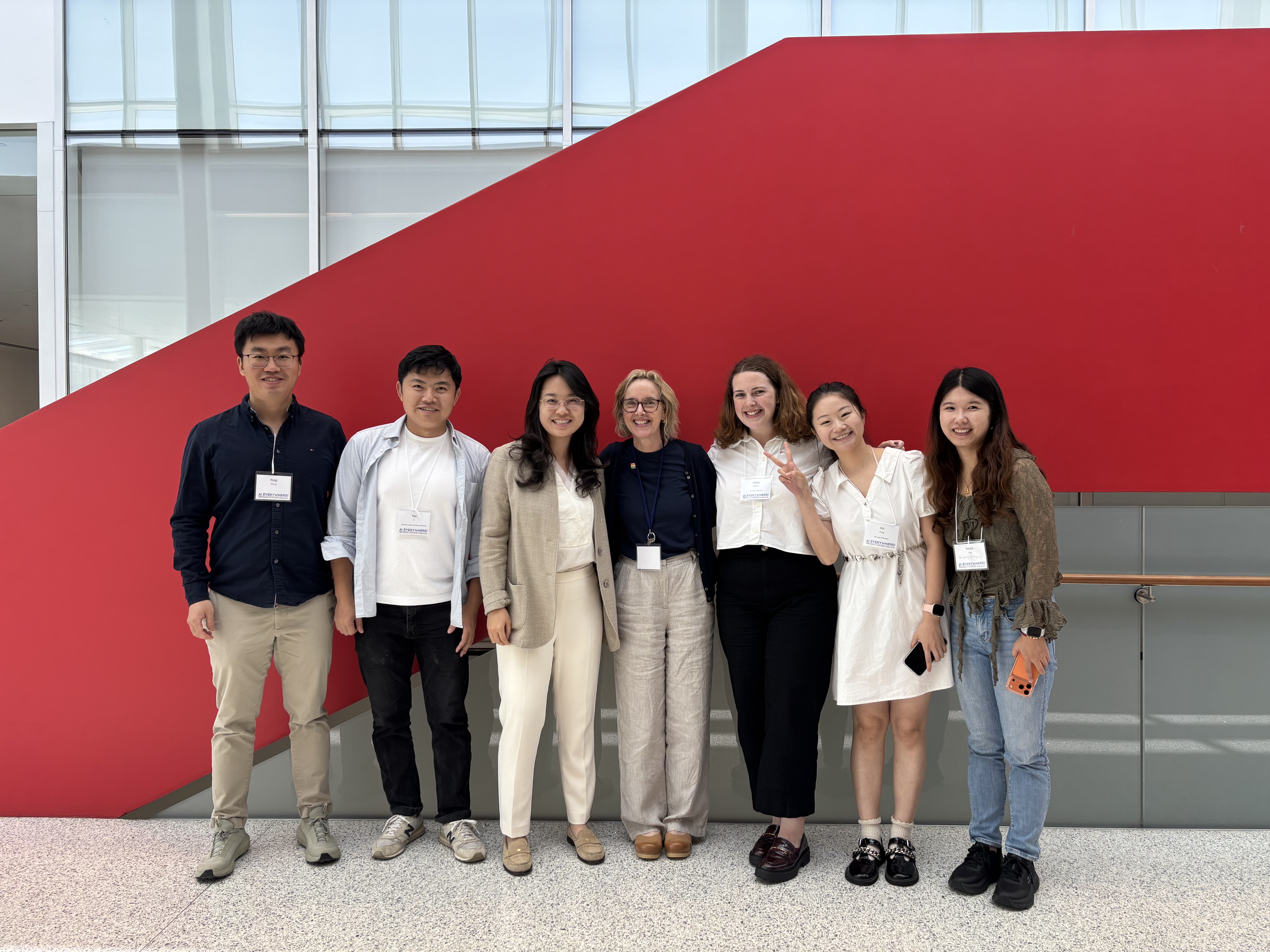 A diverse group of seven people wearing name tags pose smiling together in front of a bold, bright red architectural feature in a modern indoor atrium.