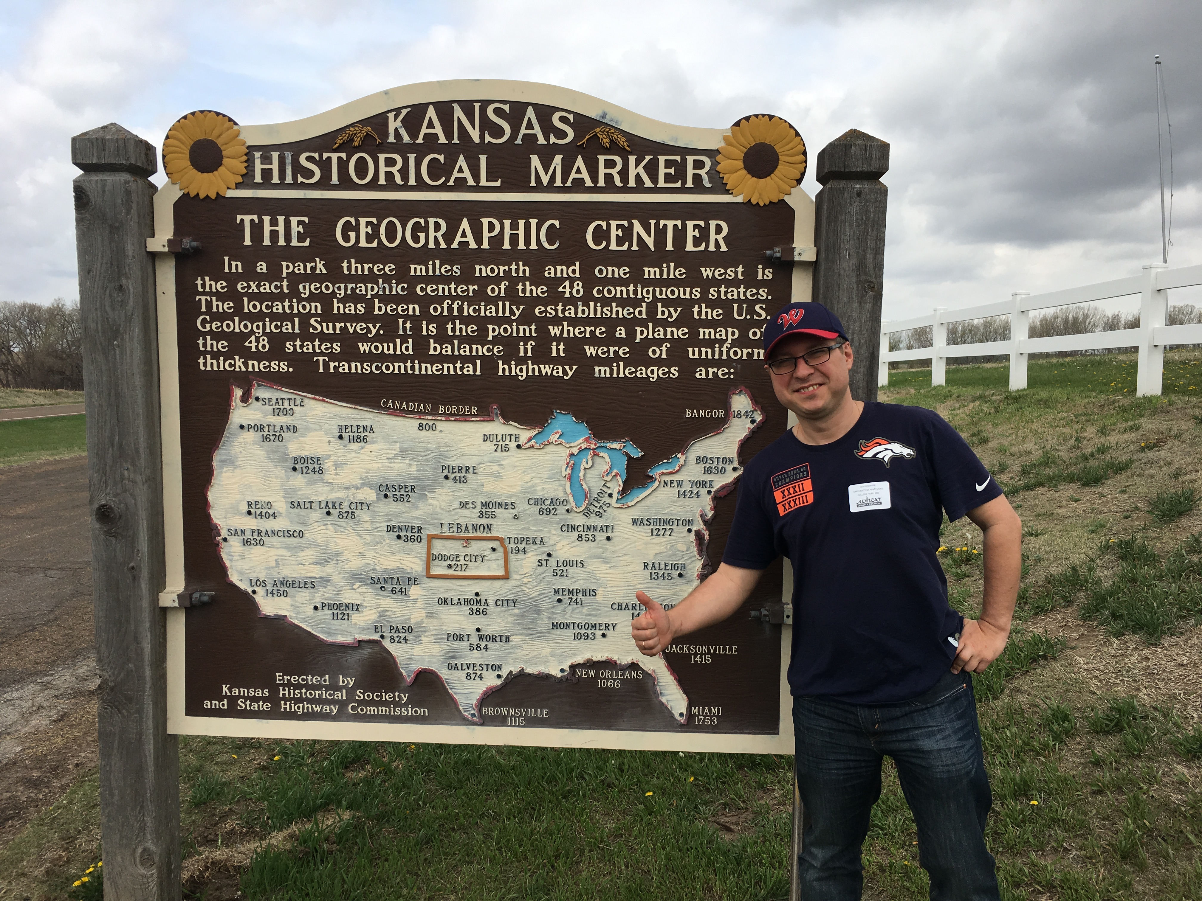 A man stands outdoors next to a large brown wooden Kansas Historical Marker sign titled 