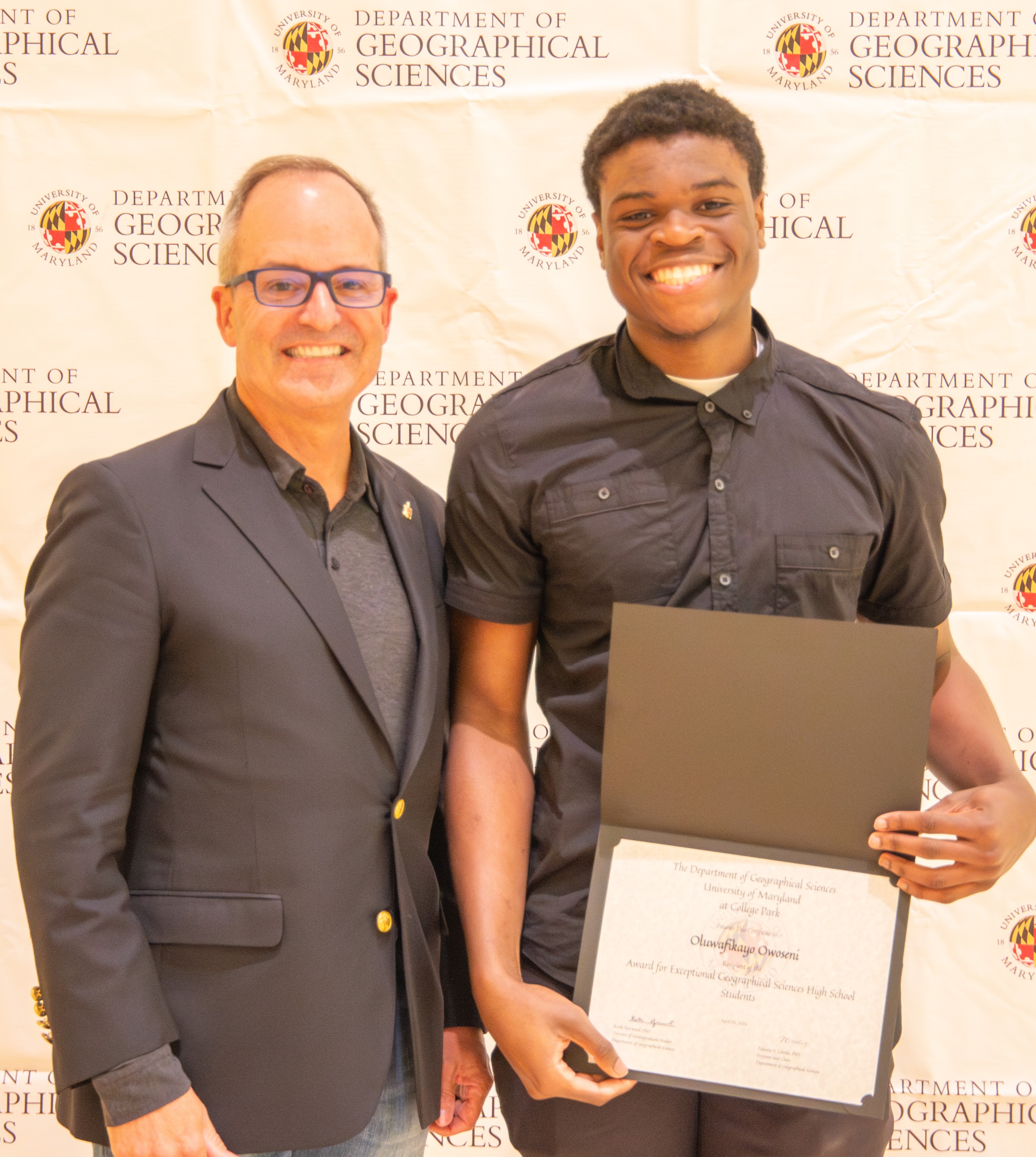 A student, Oluwafikayo Owoseni, smiles while holding an Award for Exceptional Geographical Sciences High School Students certificate alongside a faculty member at a University of Maryland event.