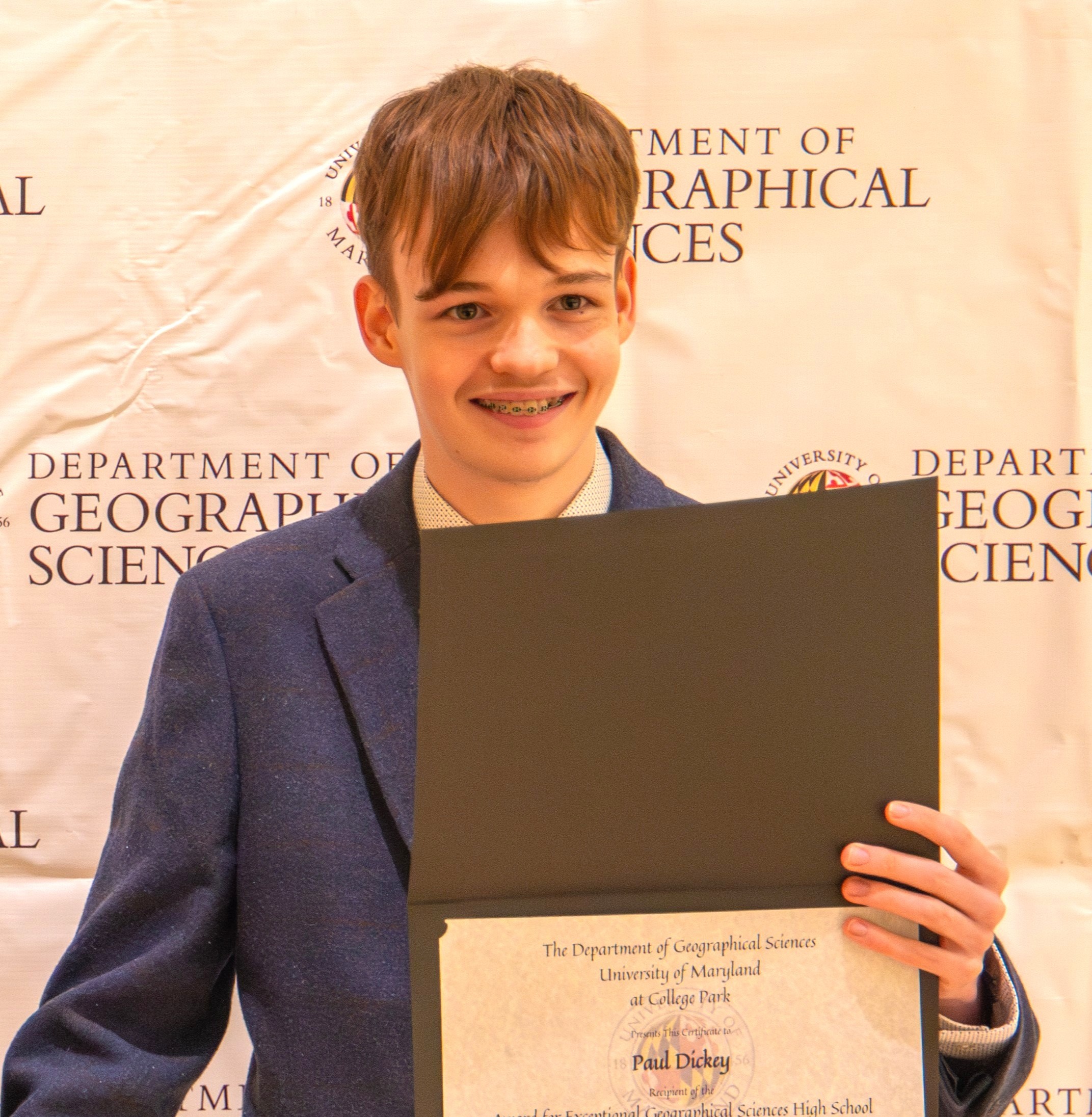 High school student Paul Dickey smiles while displaying his Award for Exceptional Geographical Sciences High School Students certificate in front of a University of Maryland Department of Geographical Sciences backdrop.