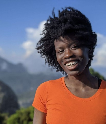 A Black woman with natural coiled hair smiling brightly in a bright orange t-shirt, set against a blurry backdrop of mountains and a blue sky.