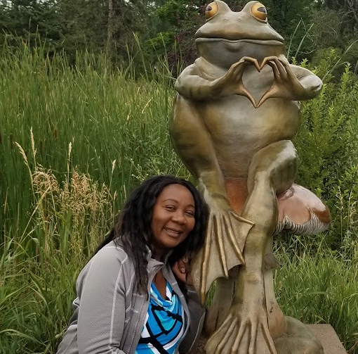 A smiling dark-skinned with long dark hair rests her head against a large frog statue outdoors. The frog is seated atop a mushroom, holding its hands together in a heart shape. Lush green foliage and tall grasses fill the background.