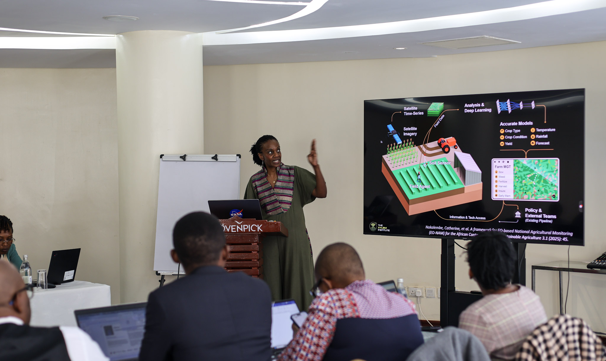 A female presenter stands by a flip chart, writing as she addresses a group of seated professionals. To her left, a large screen displays a presentation titled 