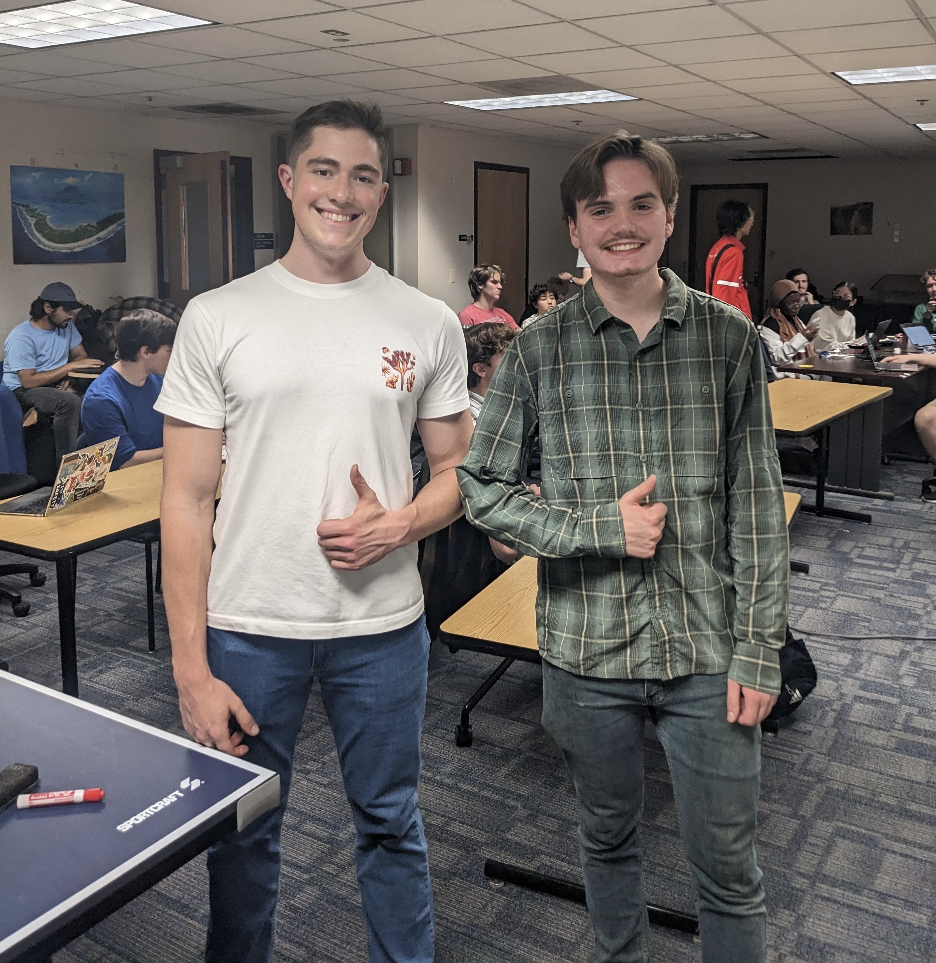 Two young men stand side by side smiling and giving thumbs-up gestures inside a brightly lit indoor common area or classroom. The man on the left wears a white t-shirt with a small graphic on the chest and blue jeans, standing next to a ping pong table. The man on the right wears a green plaid long-sleeve shirt and grey pants. In the background, other people are seated at study tables across the carpeted room.
