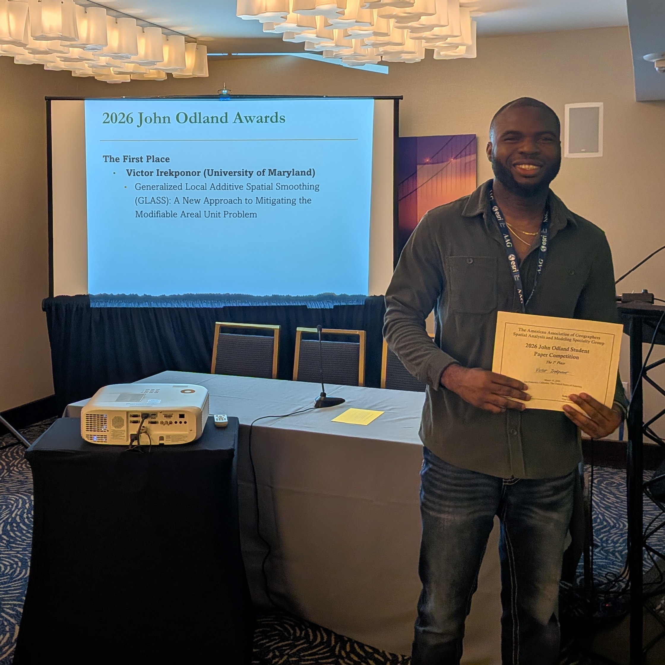 A smiling man stands in a conference room holding a certificate for the 2026 John Odland Student Paper Competition (1st Place). He is wearing a grey button-down shirt, jeans, and an ESRI/AAG lanyard. Behind him, a projector screen displays his name, Victor Irekponor (University of Maryland), and the title of his winning paper: 