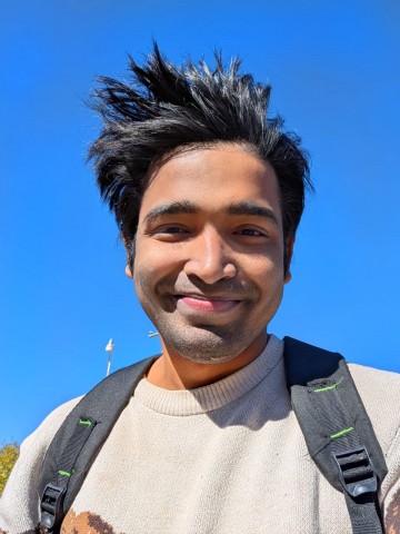 A young South Asian man smiling gently outdoors in a off-white sweater and backpack under a clear blue sky.