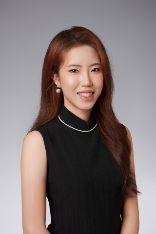A professional headshot of a young East Asian woman with long, reddish-brown hair, smiling at the camera against a seamless studio grey gradient background. She is wearing a sleeveless black high-neck top accented with a delicate white pearl trim along the collar, paired with matching pearl drop earrings.