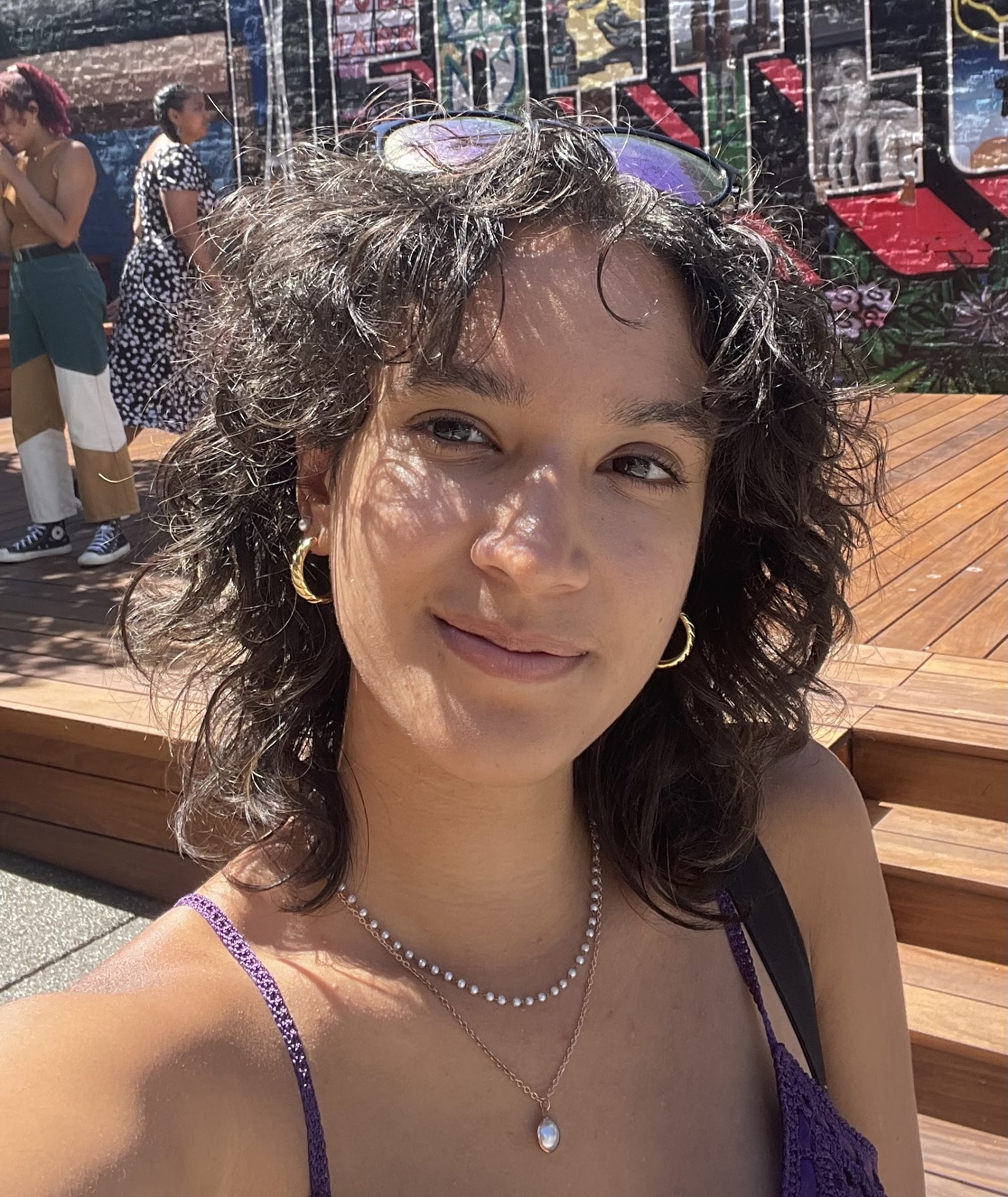 A young, light-skinned woman with dark curly hair smiling outdoors on a sunny day in a purple top.