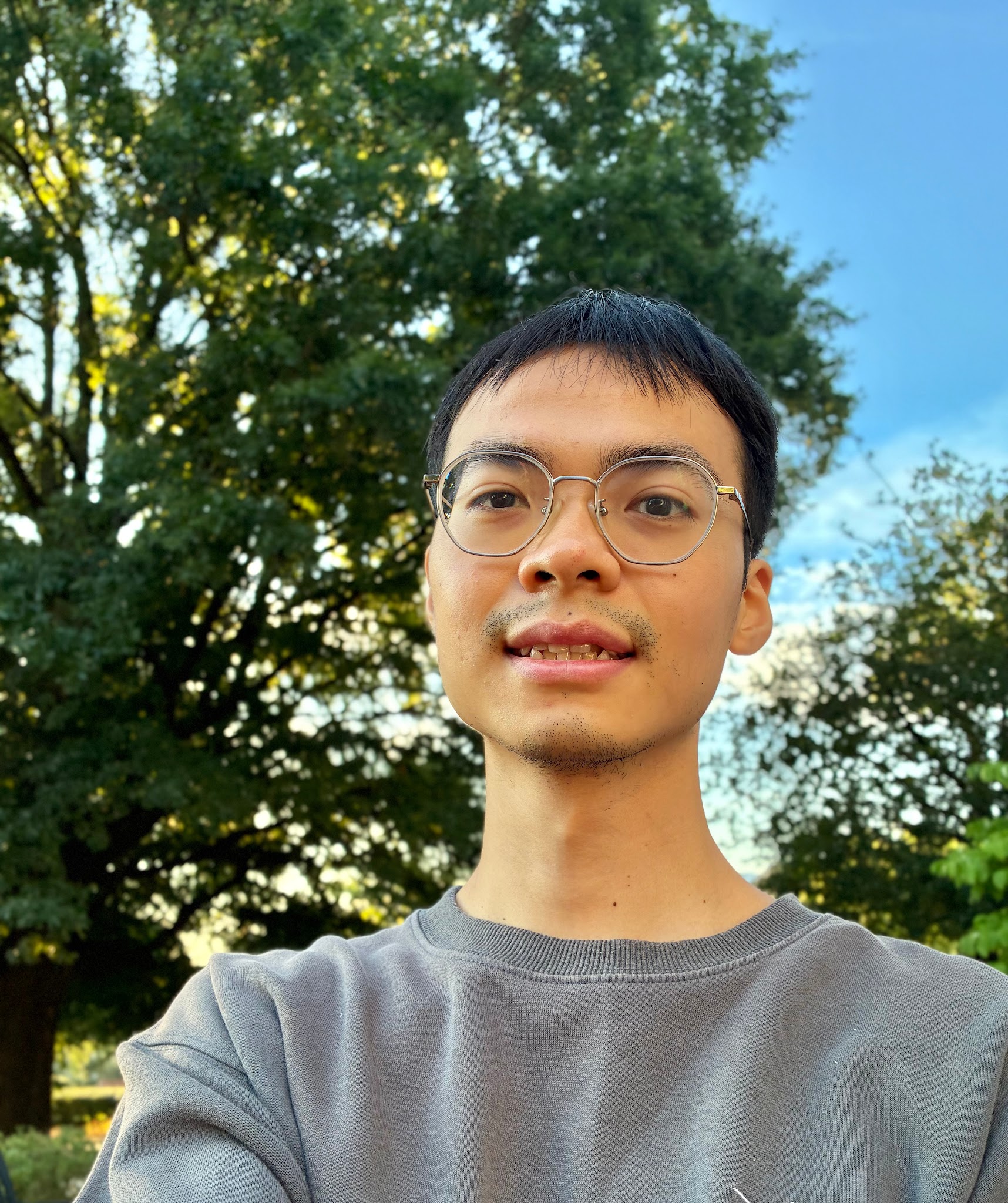 An East Asian man with round wire-rimmed glasses smiling outdoors in a gray sweatshirt against a lush green tree.