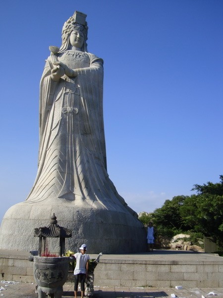 A low-angle shot of a towering stone statue of a female deity, likely Mazu, carved from light-colored granite. The figure wears an ornate, traditional crown and flowing robes, holding a ceremonial object in both hands. At the base of the massive sculpture, a person stands near a large incense burner, and another person stands near the statue’s foot, providing a sense of its immense scale against a clear blue sky.5