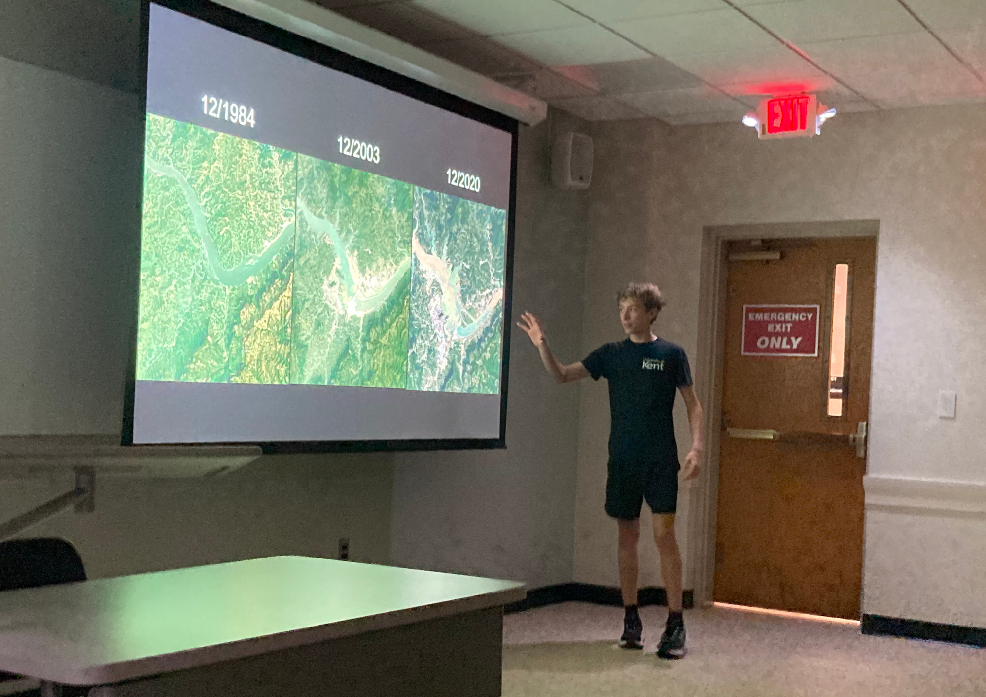 High school student Simon Mitchell stands in front of a projection screen gesturing toward three satellite images of the Three Gorges Dam region in China labeled December 1984, December 2003, and December 2020.