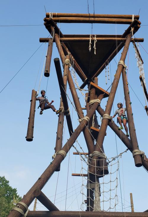 Two students in harnesses are climbing a wooden structure that is several stories tall