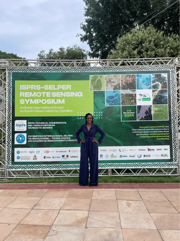 Catherine Nakalembe poses with a big smile and her hands on her hips in front of a large sign about the ISRPS-SELPER Remote Sensing Symposium.