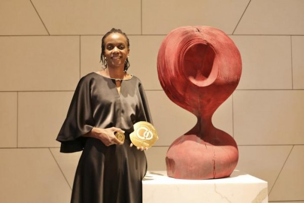 Catherine Nakalembe holds a plaque in front of a modern-looking sculpture