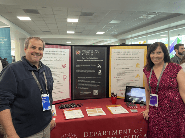 Alumn Demian Rybock and MS Program coordinator Erin Jacobs stand beside a table displaying a poster with information about the program.