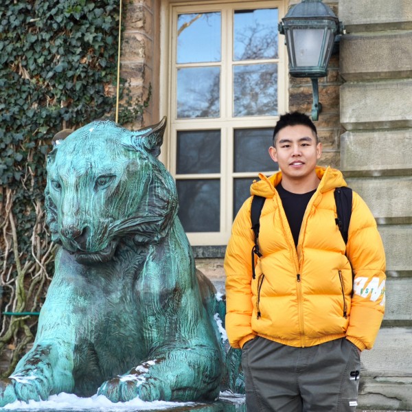 ​Xueyuan (Eric) Gao, dressed in a yellow jacket, stands next to the bronze tiger at the front of Nassau Hall at Princeton University