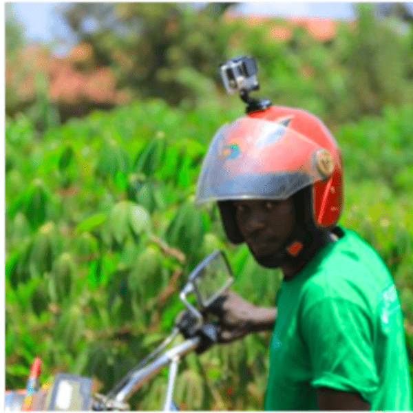 Motocyclist wears a helmet with a camera on top in front of a green field.