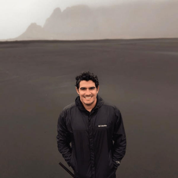 A man poses in front of a striking black sand beach, with gentle waves lapping at the shore.