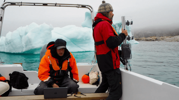 Two men covered by winter gear sitting on a boat on icy water, one working on a computer and the other about to put a device on the water
