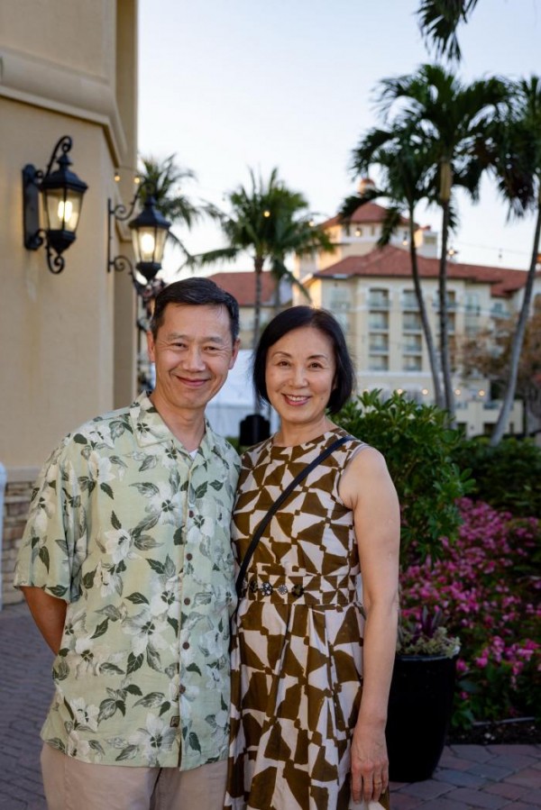 An middle age couple smiles while standing outdoors in front of a Mediterranean-style resort building with palm trees and lush plants in the background. The man wears a Hawaiian shirt and the woman a sleeveless geometric-print dress.