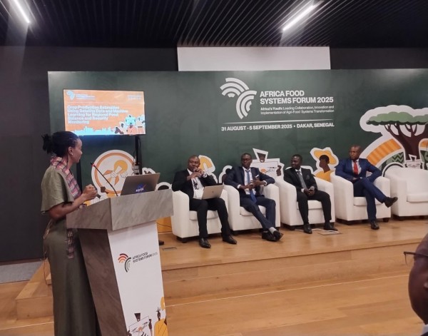 A woman presents at a podium to five seated panelists at the Africa Food Systems Forum 2025, with the conference branding and a screen about crop monitoring visible on the stage backdrop.