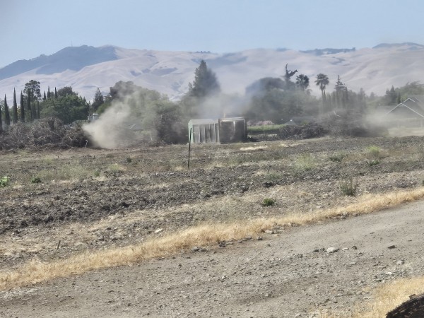 A landscape photograph shows a small structure engulfed in flames and emitting heavy smoke in the middle of a dry, tilled field. The foreground features a dirt road, while the background shows brush piles, trees, and rolling hills under a blue sky.