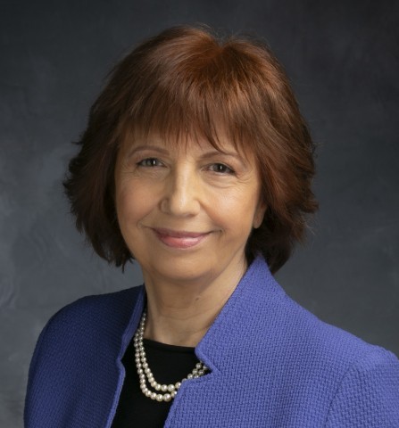 A head-and-shoulder portrait of a smiling woman with reddish-brown hair, wearing a periwinkle blue blazer and a pearl necklace against a dark grey background.