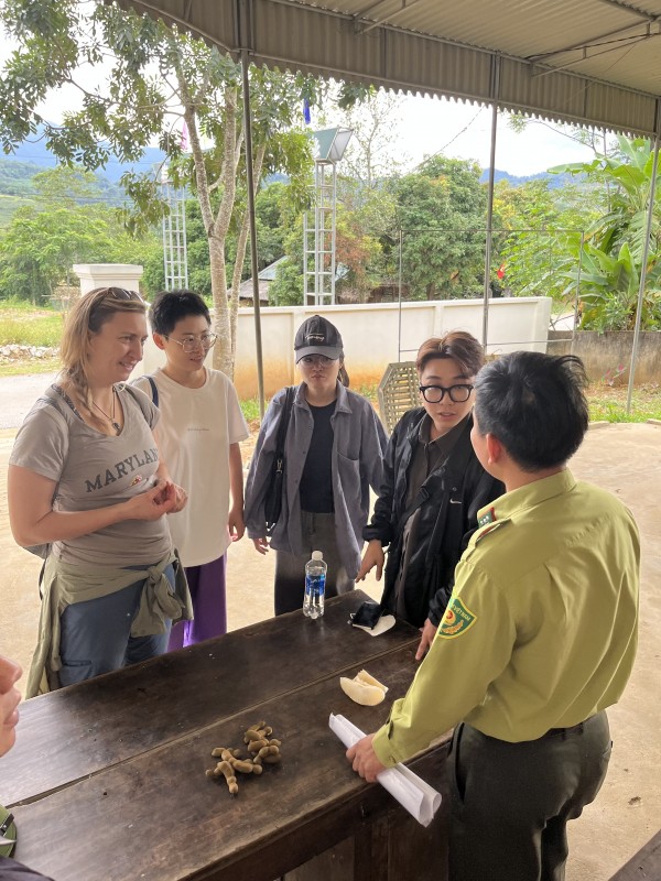 Four people stand at a wooden table outdoors in a rural, mountainous setting, engaging in a conversation with a Vietnamese official in a green uniform. On the table are tamarind pods, sliced fruit and a water bottle.