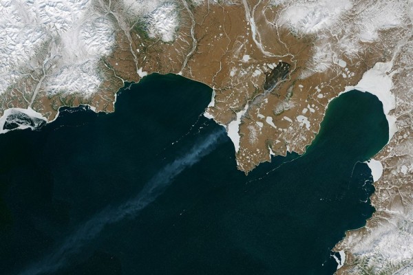 A satellite view shows a snow-dusted, brown coastline where a long, misty plume originates from a dark land scar and stretches across a deep teal sea. Bright white sea ice and frozen lakes contrast with the dark water and rugged tundra.