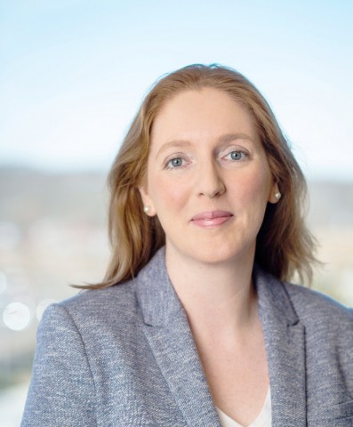 A professional headshot of a woman with strawberry-blonde hair and blue eyes, smiling warmly while wearing a blue textured blazer and pearl earrings against a soft, blurred outdoor background.