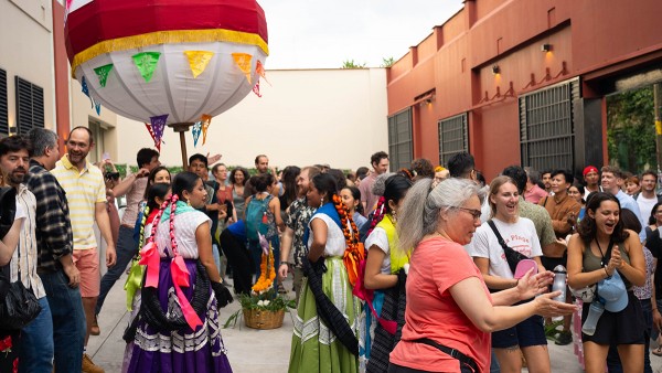 A large, diverse group of people gathers in an outdoor courtyard in Oaxaca, Mexico, for a lively social event. In the foreground, two women in traditional Oaxacan dress—vibrant purple and green skirts with intricate white lace and ribbons braided into their hair—stand near a large, white and red decorative ceremonial balloon (marmota) adorned with colorful papel picado flags. Around them, many other attendees in casual attire are smiling, talking, and dancing. The background features warm, terracotta-colore