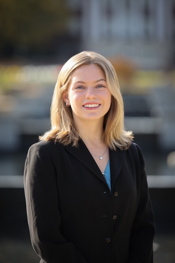 A medium close-up headshot of a young woman with shoulder-length, wavy, dirty blonde hair and blue eyes, who is smiling broadly and looking at the camera. She is standing outdoors on an overcast day, in a blurred background that shows trees and buildings, with her arms crossed at the waist. Her black blazer with two buttons is zipped up and she is wearing a thin silver chain with a small heart pendant.