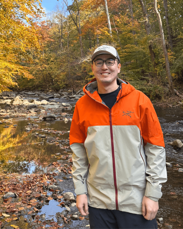A smiling man stands in front of a shallow, rocky creek surrounded by a forest with vibrant yellow and orange autumn foliage. He is wearing a grey baseball cap, glasses, and a two-tone Arc'teryx outdoor jacket that is bright orange on the top half and light grey on the bottom. The ground is covered in fallen autumn leaves, and the trees are reflected in the calm water.