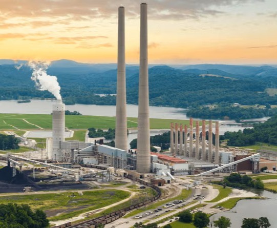 Aerial wide shot of a large coal-fired power plant nestled in a lush, mountainous valley. Two massive, slender concrete chimneys dominate the center, flanked by a shorter white cooling tower emitting a plume of steam. To the left, a large pile of coal sits near conveyor belts and rail lines, while a river curves around the facility in the foreground, all set against a backdrop of rolling green hills and a hazy blue mountain range under a warm, sunset-lit sky.