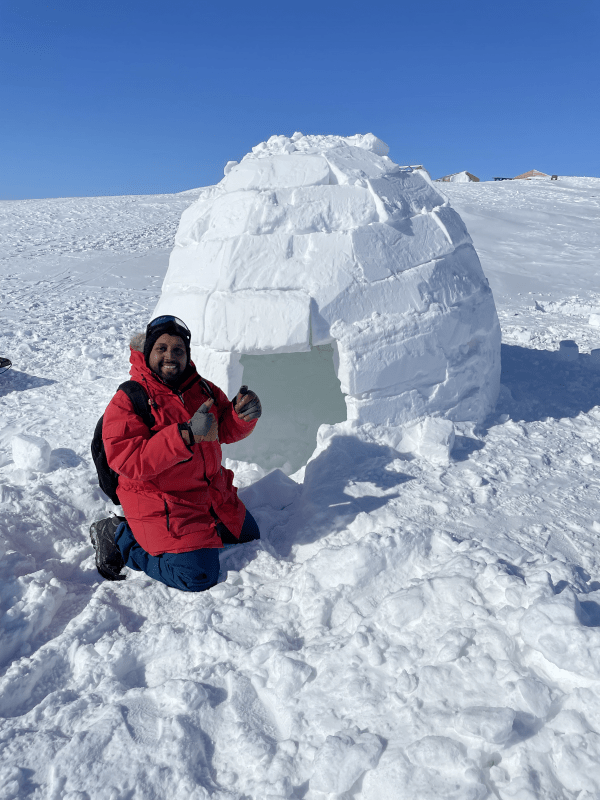 A man in a bright red parka, blue snow pants, and a backpack kneels in the snow next to a large, hand-built igloo made of stacked snow blocks. He is smiling and giving a thumbs-up. The scene is set on a vast, sunlit snowfield under a clear blue sky, with a few distant structures visible on the horizon.