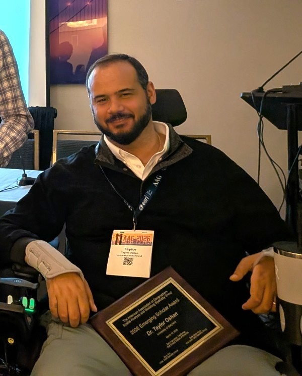 A medium shot of a man with a beard, wearing a black quarter-zip sweater and an AAG 2026 conference badge, smiling and holding a wooden plaque. The plaque reads, "The American Association of Geographers Spatial Analysis and Modeling Specialty Group, 2026 Emerging Scholar Award, Dr. Taylor Oshan, University of Maryland." He is seated in a wheelchair in what appears to be a conference room.