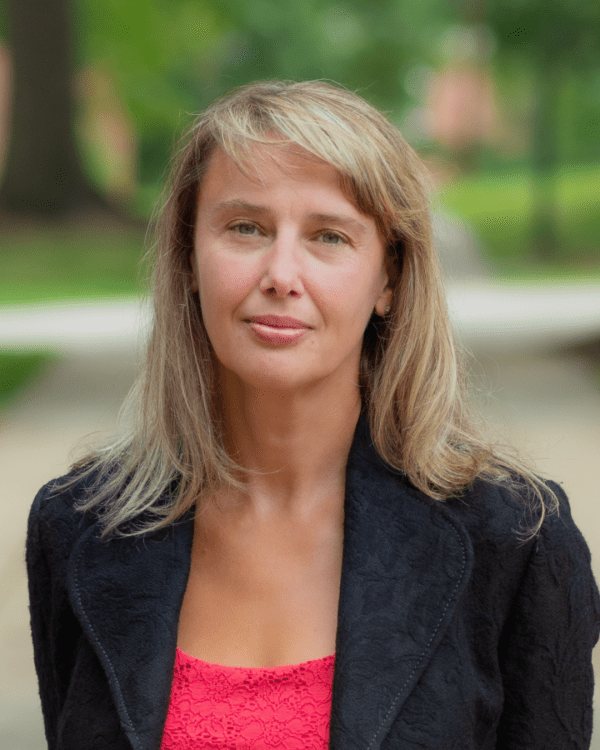 A medium headshot of a woman with light brown hair in a black textured blazer and red top, smiling slightly in a blurred outdoor setting.